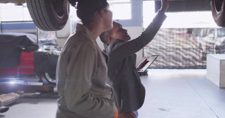 Women in mechanic coverall, blazer discussing inspection of lifted car in garage, with lift, tablet. Automotive, teamwork, industry, vehicle maintenance, professional, industrial, collaborationの写真素材