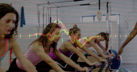 Pedaling six women in athletic wear on bikes in gym, with microphone boom, blue exercise ball....