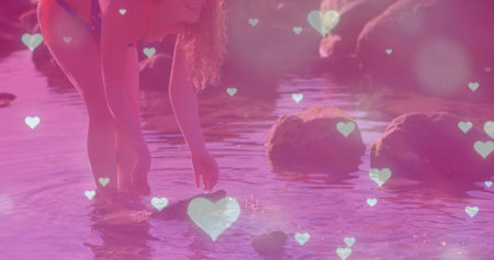 Bending woman wearing red swimsuit touching water at rocky beach, with heart overlays, copy space. Nature, serenity, relaxation, romantic, playful, scenic, leisureの写真素材