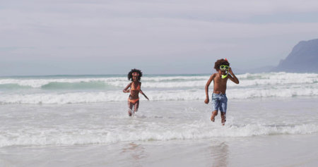 Running siblings in orange and blue swimwear through waves at beach, with swim goggles, snorkel set. Playful, adventure, youth, seaside, leisure, exploration, carefreeの写真素材
