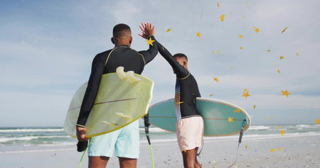 High-fiving surfers holding surfboards with leashes on sandy beach facing ocean under cloudy sky....