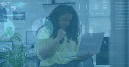 Sitting woman wearing work attire holding laptop and resting chin in office with monitors, plants. Professional, workspace, contemporary, technology, collaboration, productivity, minimalistの写真素材