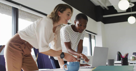 Collaborating adult women pointing at laptop screen in modern office, with coffee mug notebook. Professional, teamwork, modern, office, communication, productivity, workspaceの写真素材