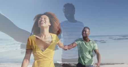 Walking African American couple in yellow dress holding hands along sand near rocky coast, smiling....
