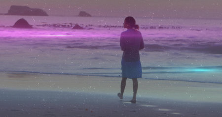 Standing barefoot woman in blue dress facing sea on beach at dusk, purple-blue band and rocks. Serenity, tranquility, reflection, nature, minimalism, contemplative, oceanの写真素材