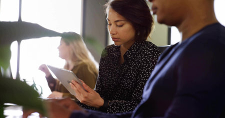 Reading woman in black blouse holding tablet at bright office, with laptop, plant, handheld device. Collaboration, productivity, modern, minimalistic, technology, workspace, wellnessの写真素材