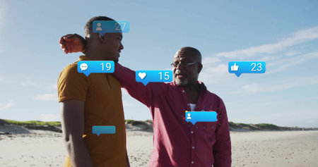 Talking father in red shirt guiding son in yellow t-shirt on beach with social media notifications. Mentorship, generational, connection, outdoor, vibrant, communication, leisureの写真素材