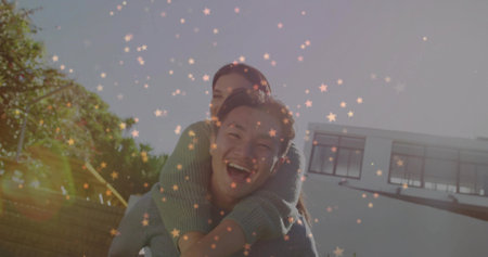 Laughing Korean man giving piggyback ride to woman in modern white house backyard, with lens flares. Joy, friendship, outdoor, leisure, modern, casual, vibrantの写真素材