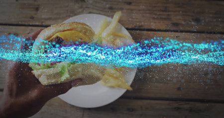Lifting sesame-seed bun burger with adult hand over wooden tabletop, with ceramic plate of fries....