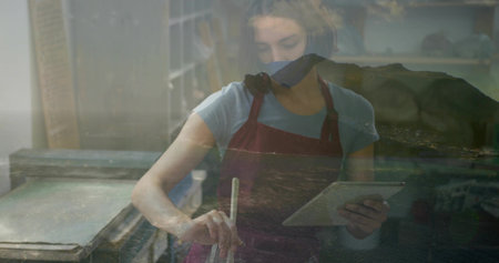 Stirring bowl mixture artisan wearing red apron at studio worktable, with tablet and mixing stick. Creative, craft, artisan, workspace, modern, diverse, productivityの写真素材