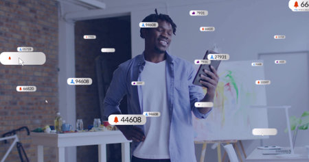 Smiling man wearing denim shirt holding smartphone and showing notifications in studio, with canvas. Artistic, creative, modern, vibrant, inspiring, digital, workspaceの写真素材