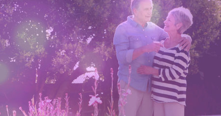 Basking senior couple standing embracing in garden at golden hour, with ceramic mug and flowers. Romance, tranquility, vitality, nature, relaxation, nostalgia, harmonyの写真素材