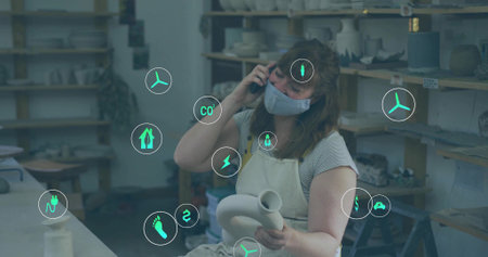 Holding clay vessel, apron-clad ceramic artist talking on smartphone at workshop. Craftsmanship, sustainability, artistry, studio, handmade, eco-friendly, creativeの写真素材