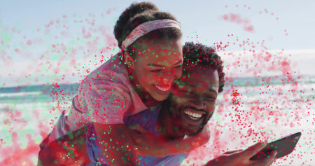 Smiling couple taking piggyback ride at sunlit sandy beach, with smartphone and digital speckles. Joy, leisure, friendship, vibrancy, outdoor, relaxation, affectionの写真素材