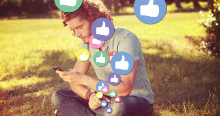 Sitting man wearing dotted shirt scrolling smartphone in park, with floating thumbs up icons....
