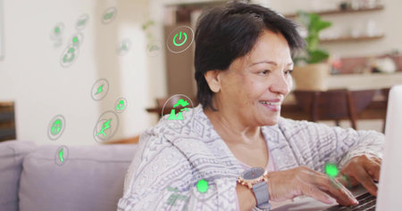 Typing senior Indian woman sitting on sofa in living room, with laptop, eco icons, copy space....