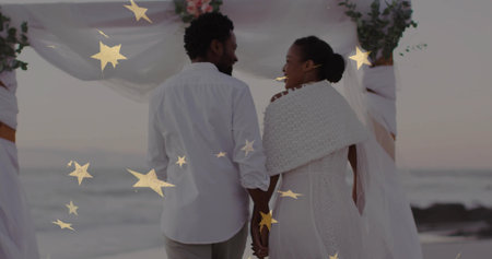Vowing couple wearing wedding attire on beach at sunset, with draped arch, gold stars, waves. Romance, celebration, elegance, serenity, natural, outdoor, pastelの写真素材