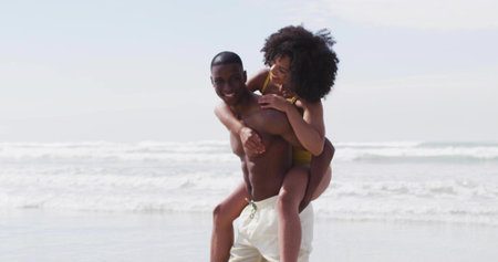 Man carrying woman on back in shallow ocean water at beach, with swim trunks, yellow swimsuit. Romance, outdoor, adventure, leisure, exhilaration, vibrant, lifestyleの写真素材