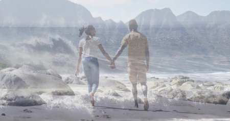 Walking couple holding hands toward water on sandy beach with rocks, waves breaking at feet....