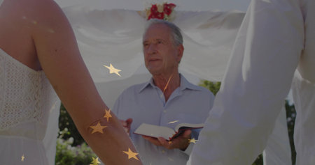 Conducting officiant reading ceremonial book under white draped arch in garden, with joined hands....