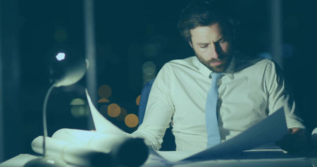 Reviewing man wearing shirt, tie at office at night with desk lamp, rolled plans, copy space. Professional, workspace, urban, nighttime, concentration, modern, skyscraperの写真素材