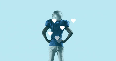 Football player standing hands on hips on light blue backdrop, wearing helmet pads. Athlete, sports,...