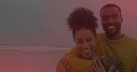 Hugging couple wearing matching mustard sweaters at beach shoreline, ocean waves under cloudy sky....