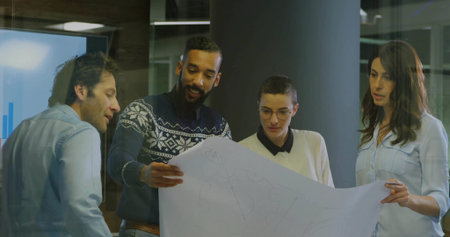 Group of professionals examining blueprint in office area, with digital charts and glass partitions....