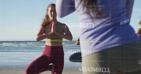 Practicing women wearing sportswear holding tree pose on shoreline, with wet sand and ocean waves....