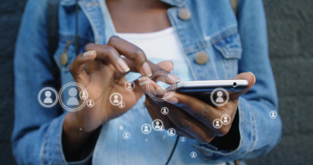 Tapping smartphone woman wearing denim jacket and white tank scrolling user icons at urban wall. Technology, connectivity, communication, social networking, urban, modern, casualの写真素材
