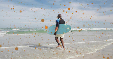 Walking senior surfer carrying blue surfboard into shallow shoreline with leash and orange spots. Ocean, shoreline, leisure, serenity, aquatic, adventure, vibrantの写真素材