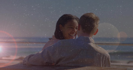 Laughing couple wearing pink and gray shirts hugging on bench at beach, with ocean, lens flare. Romance, leisure, scenic, outdoor, friendship, serenity, warmthの写真素材