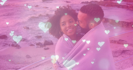 Snuggling couple sharing light-colored blanket on rocky seaside railing, with heart-shaped overlay. Romance, intimacy, serenity, affection, coastal, outdoor, tenderの写真素材