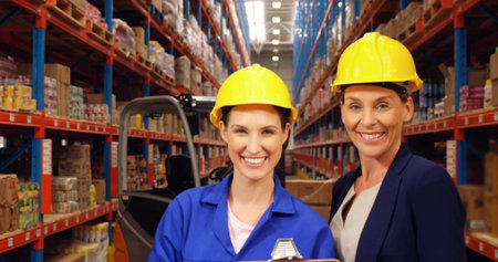 Smiling warehouse staff standing in warehouse aisle, wearing hard hats, with forklift and clipboard. Industrial, logistics, teamwork, safety, efficiency, corporate, modernの写真素材