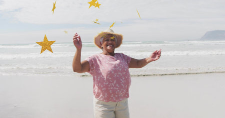 Smiling woman raising arms at beach, with straw sunhat pink floral blouse. Joy, relaxation,...