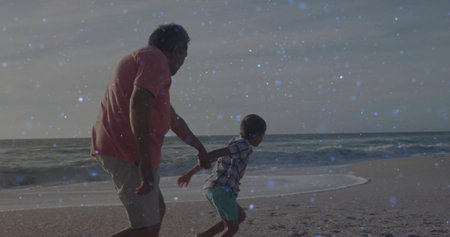 Grandfather and grandson running along sandy beach at dusk, with coral t-shirt and plaid shirt. Family, bonding, outdoor, leisure, natural, vibrant, joyfulの写真素材