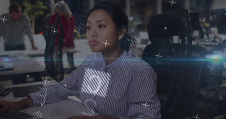 Typing mid adult Asian woman focusing on desktop monitor in open-plan office, with digital overlays....