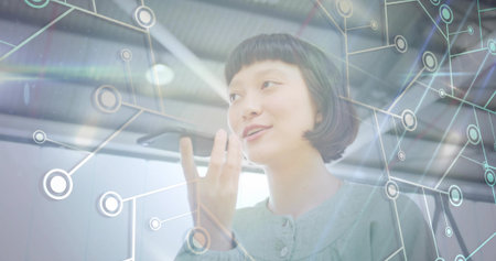 Speaking woman wearing blouse holding smartphone near mouth in office, network overlay, copy space. Technology, innovation, communication, futuristic, digital, professional, networkingの写真素材