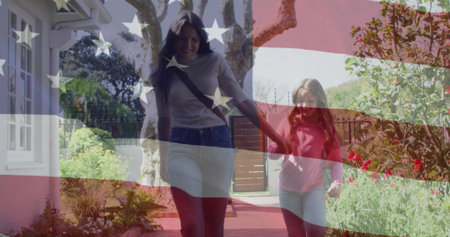 Walking woman and girl holding hands casually dressed along paved garden path, with crossbody bag....