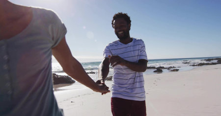 Walking couple wearing vacation clothes and holding hands on sandy beach at low tide, sunlit scene. Romance, adventure, serenity, leisure, picturesque, coastal, carefreeの写真素材