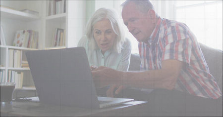Leaning forward senior couple examining laptop screen in living room with dark drink and bookshelf....