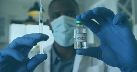 Masked male worker wearing lab coat, gloves holding syringe and vial in lab with COVID-19 injection....