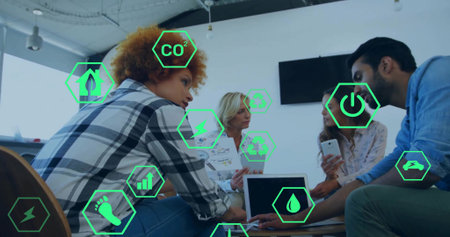 Collaborating professionals viewing phone and floating green icons around laptop in meeting room. Collaboration, teamwork, innovation, sustainability, digital, modern, workplaceの写真素材