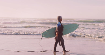 Walking senior woman carrying light green surfboard along sandy shoreline, with neoprene wetsuit....