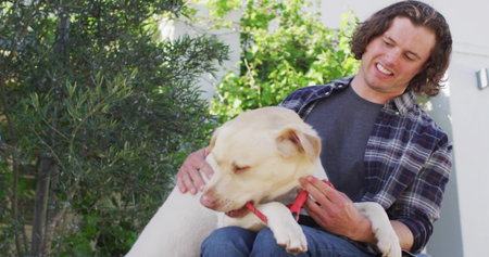 Smiling man cradling cream-colored dog on bench in backyard, with red collar and green shrubs. Pet,...