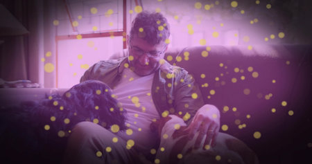 Man wearing shirt, khaki pants petting two dogs on sofa at home, with yellow bokeh overlay. Casual,...