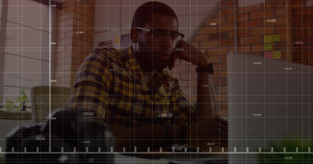 Man wearing glasses, plaid shirt resting chin, gazing at open laptop in office with camera lens. Corporate, workspace, modern, professional, productivity, brainstorming, interiorの写真素材