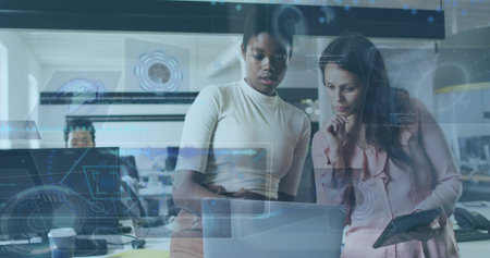 Collaborating women in work attire examining laptop in office with monitors, tablet and UI overlays. Technology, teamwork, innovation, modern, corporate, digital, communicationの写真素材