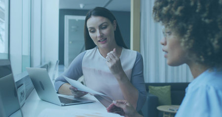 Woman in pastel blouse reviewing printed documents at modern office desk, with laptop. Professional, collaboration, workspace, modernity, teamwork, elegance, communicationの写真素材