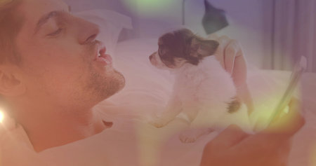 Man balancing small puppy on bed while holding smartphone in bedroom with pillows, lamp, copy space....